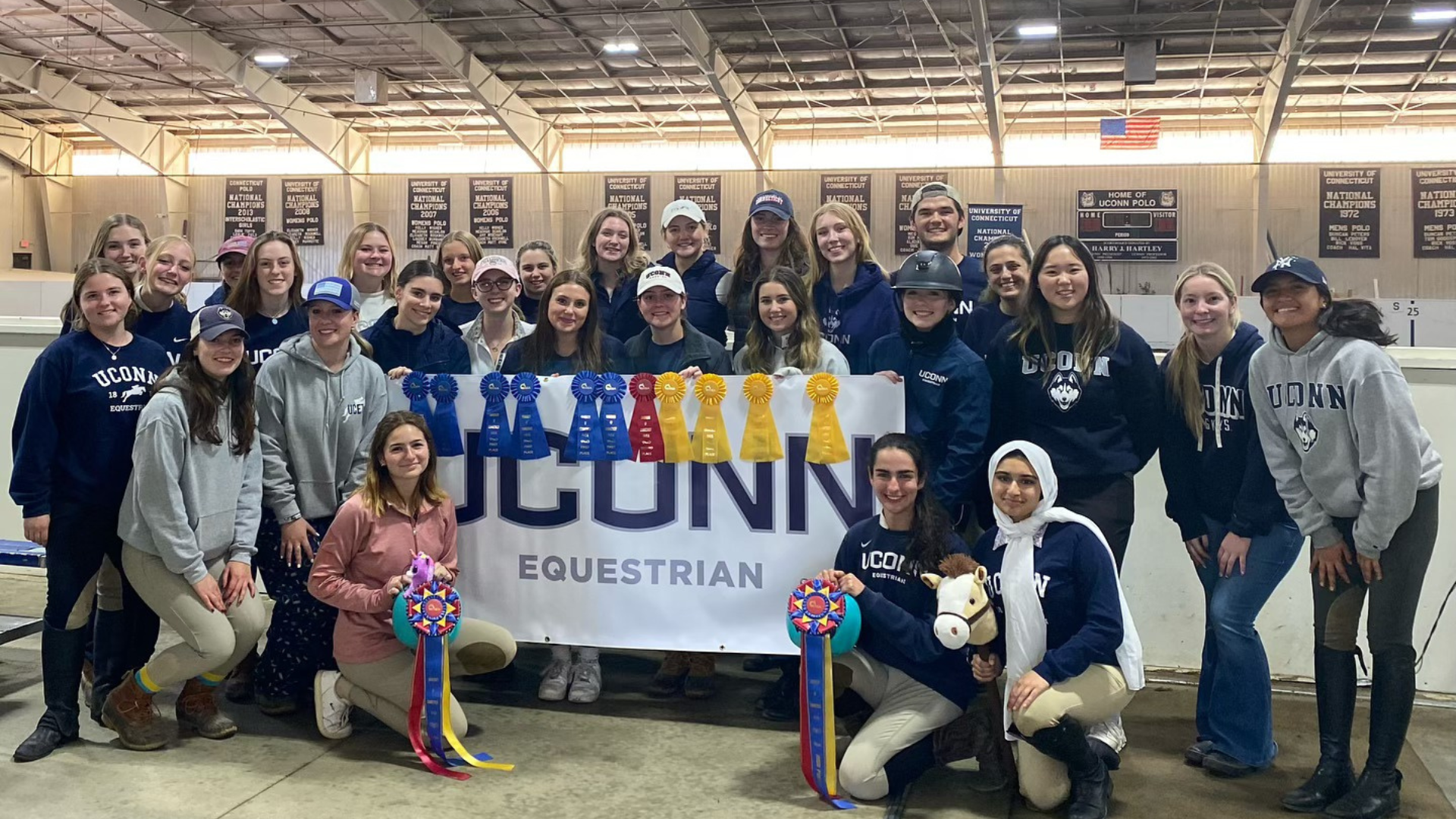 Equestrian Team posing for photo inside of Horsebarn Hill Arena with a large banner and ribbons.