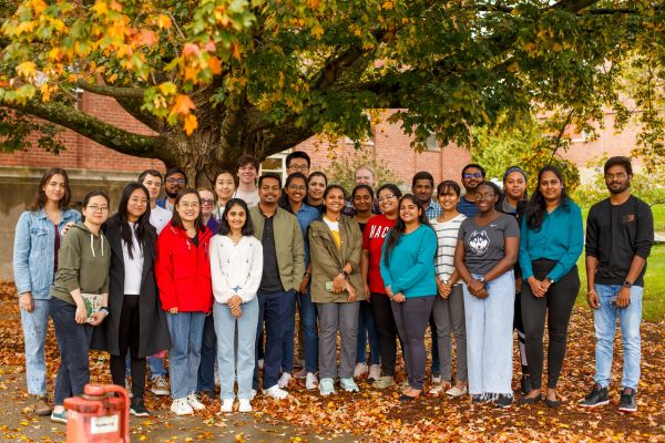 Group of graduate students posing together outside in front of a tree with red and orange leaves underneath their feet