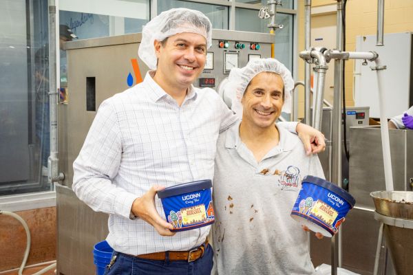 Two males standing next to each other holding tubs of ice cream in front of a machine inside of a food processing plant