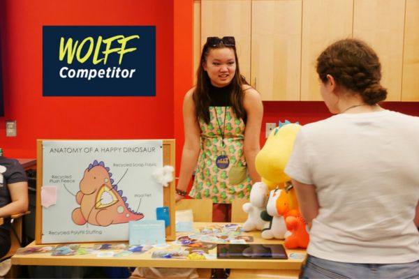 A woman stands beside a table, smiling at a colorful stuffed animal displayed on it.